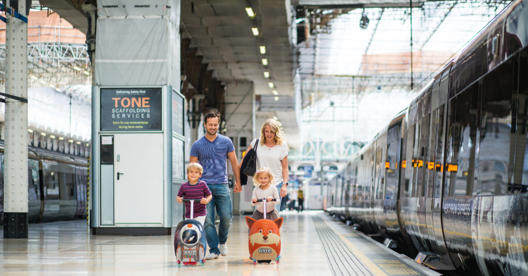 Two children in a London train station riding their luggage scooters along the train platform.