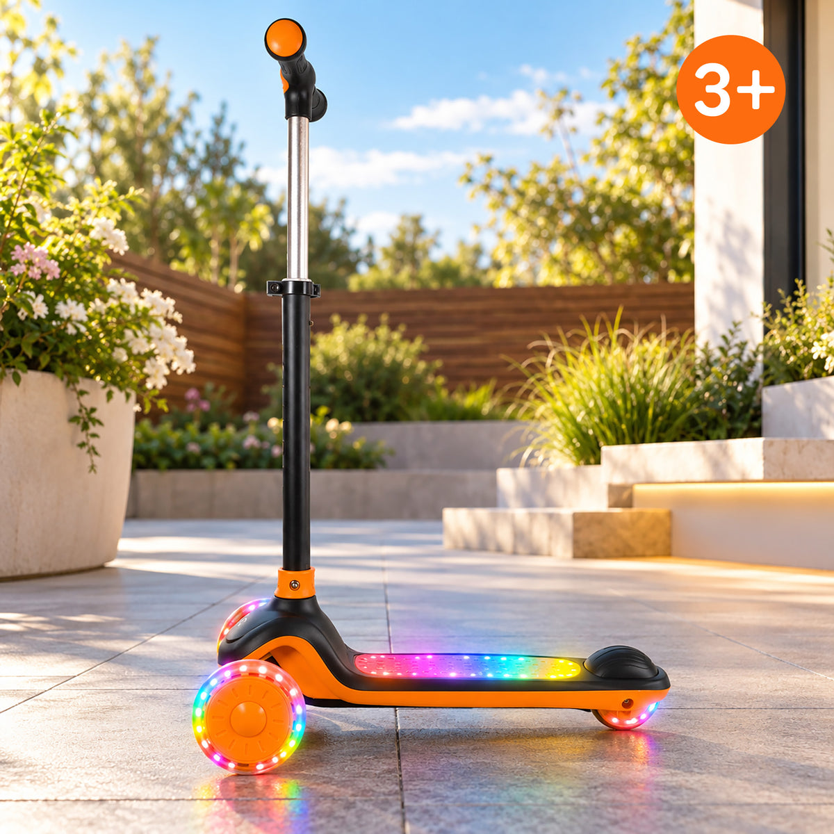 A three-wheel kick scooter with a black frame and bright orange wheels, featuring multicolor LED lights on the deck and wheels, set on a tiled patio in a sunny garden environment.