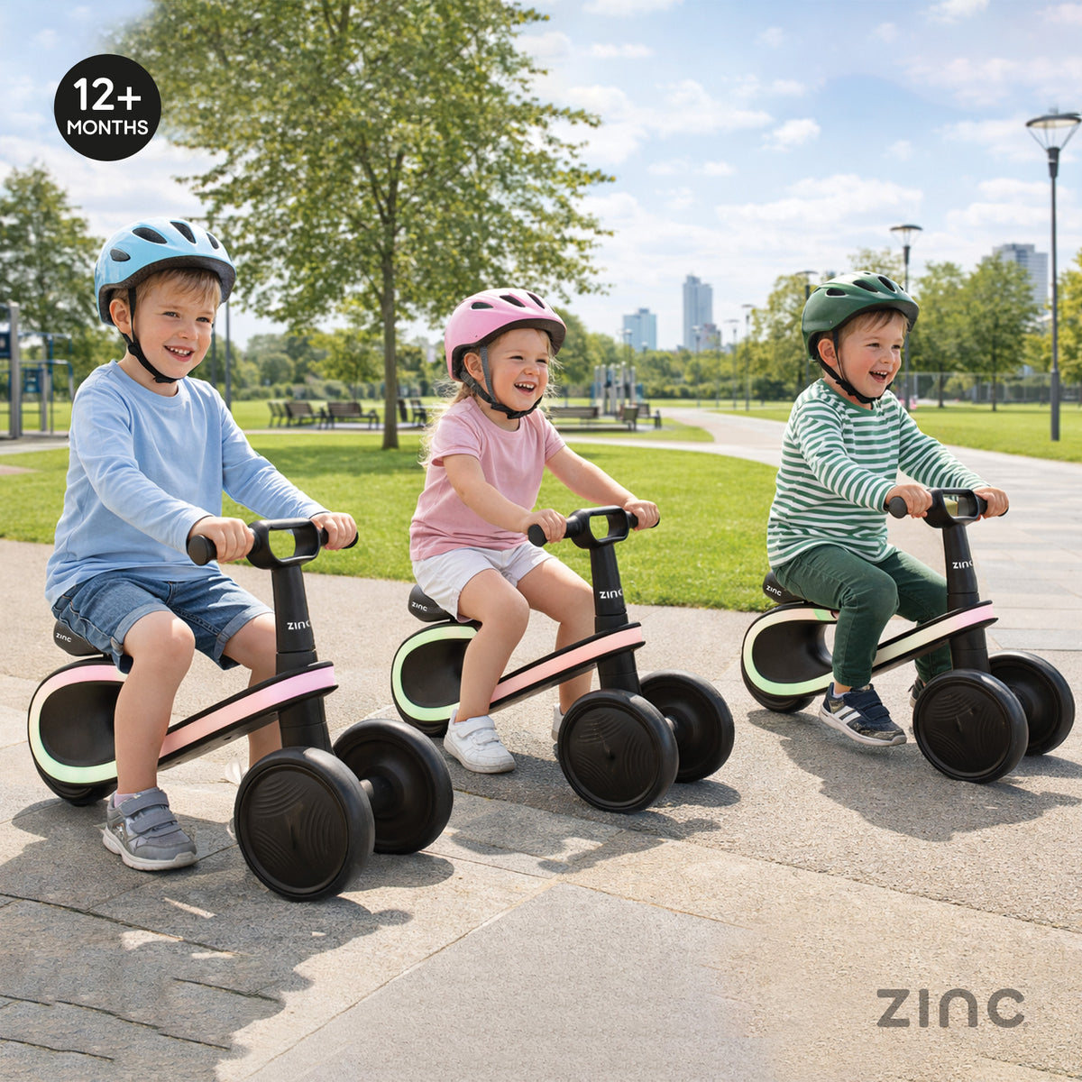 Three children wearing helmets ride colorful balance bikes on a paved path in a park. The bikes have black wheels and handles, with pink, green, and blue frame accents.