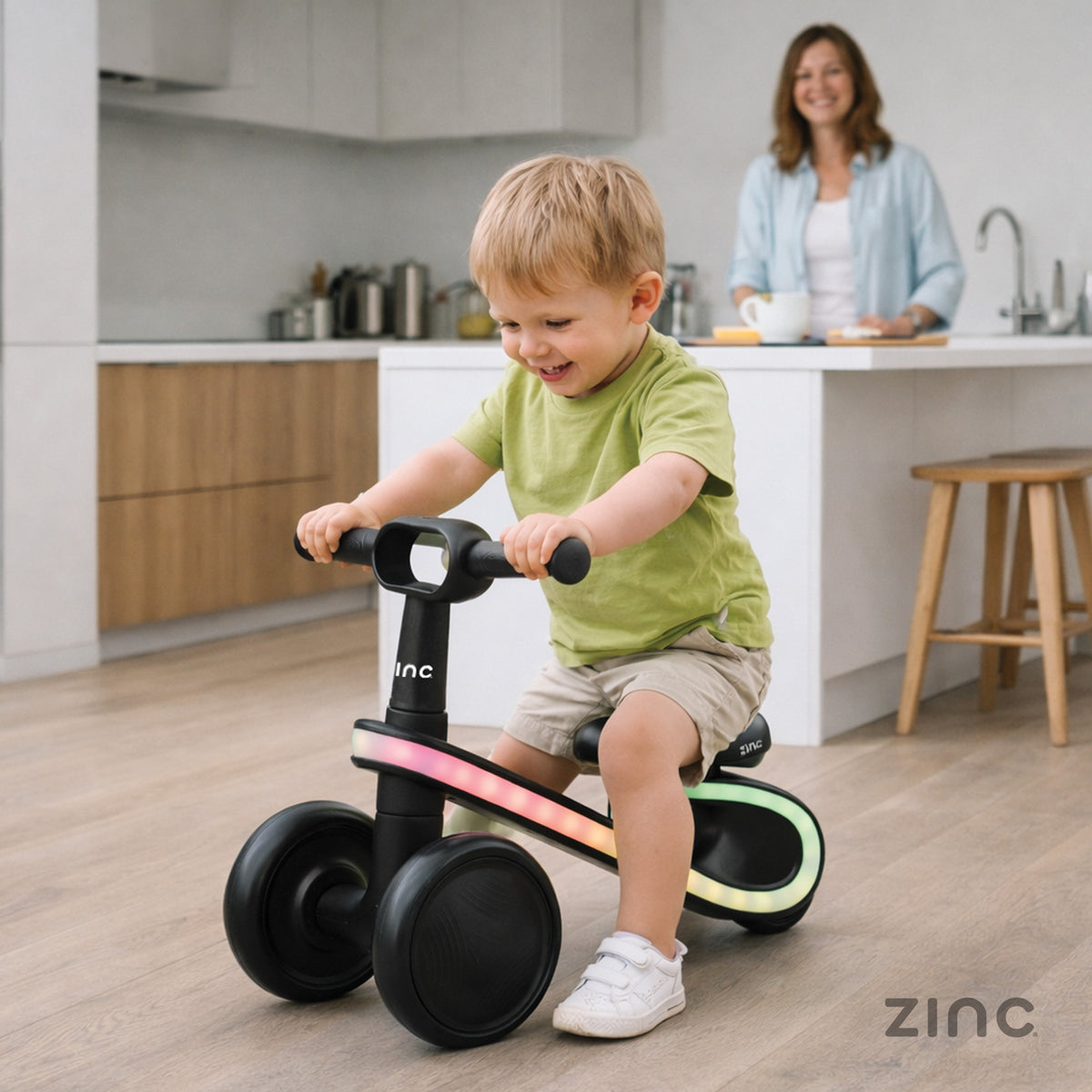A young child riding a black balance bike with colorful LED light strips in a bright modern kitchen. The brand name 'zinc' is visible on the bike frame and as a watermark in the bottom right corner.