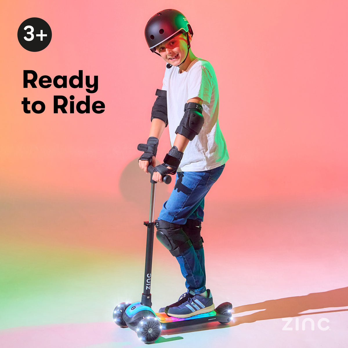 A child wearing a black helmet, white t-shirt, and blue jeans rides a black and blue Zinc scooter with glowing wheels on a pink and orange gradient background. Text on the left reads '3+ Ready to Ride'.