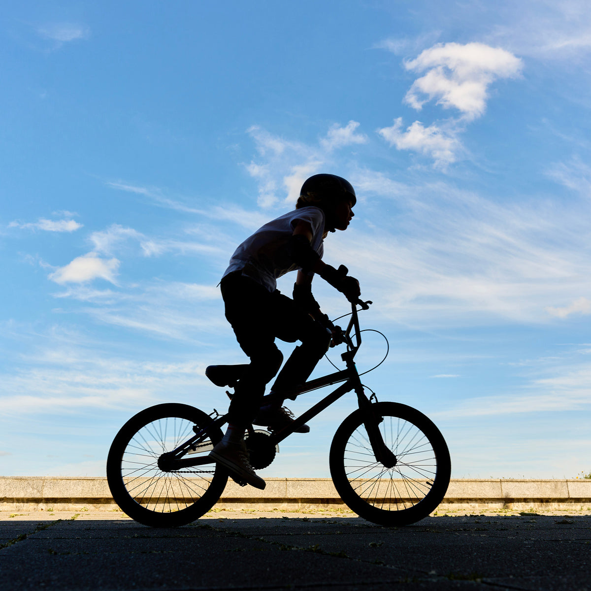 A child riding a bicycle in silhouette against a bright blue sky with light clouds.