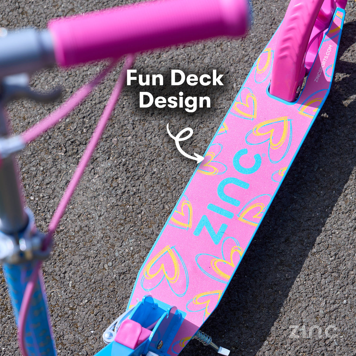 A close-up outdoor shot of a pink and blue zinc scooter deck with a heart pattern, featuring the brand name 'ZINC' on the deck and a pink handlebar.