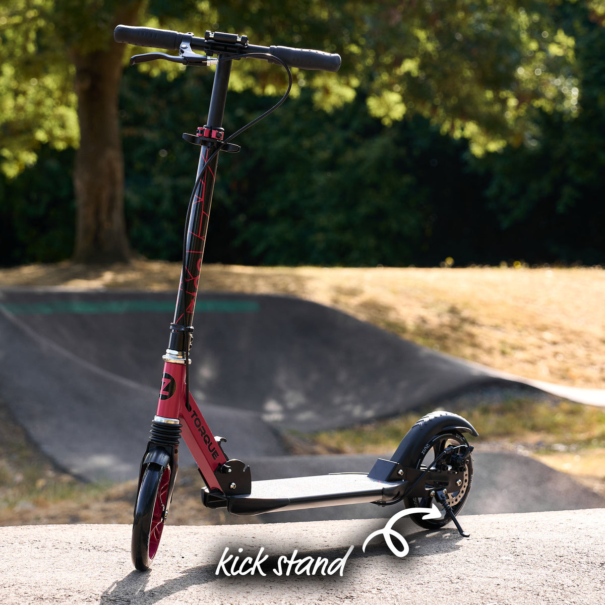 A red and black kick scooter with a black handlebar and deck stands on a concrete surface in an outdoor park setting. A white 'kick stand' logo overlay is visible at the bottom.