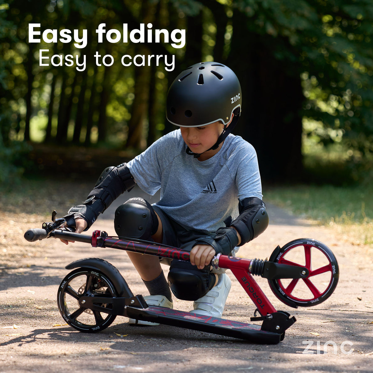 A child wearing a black helmet and protective gear kneels on a paved path outdoors, demonstrating a folding scooter. The scooter has a black deck and frame with red accents on the wheel spokes and handlebar.