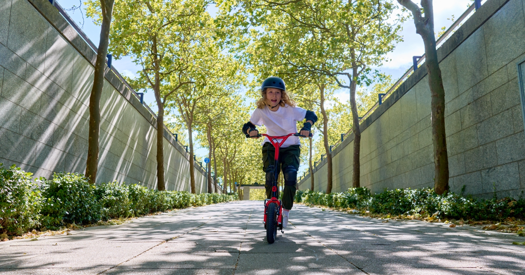 Zinc Radikal BMX Scooter in Orange being ridden down a grey path by a young boy with trees lining each side of the path