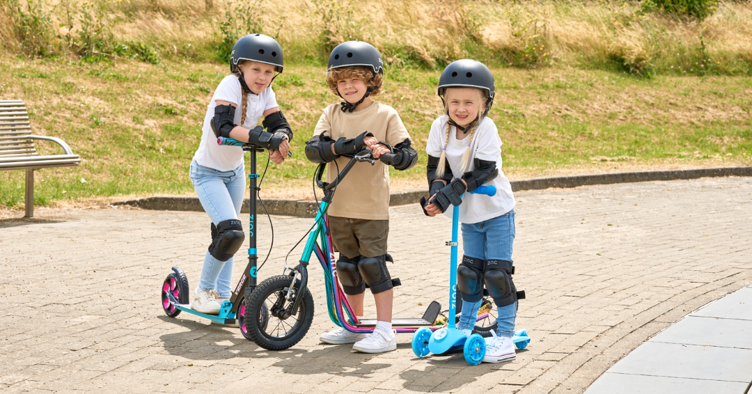 3 children with their zinc scooters, big wheel scooter, bmx scooter and 3 wheel scooter. All kids wearing helmets and safety gear.
