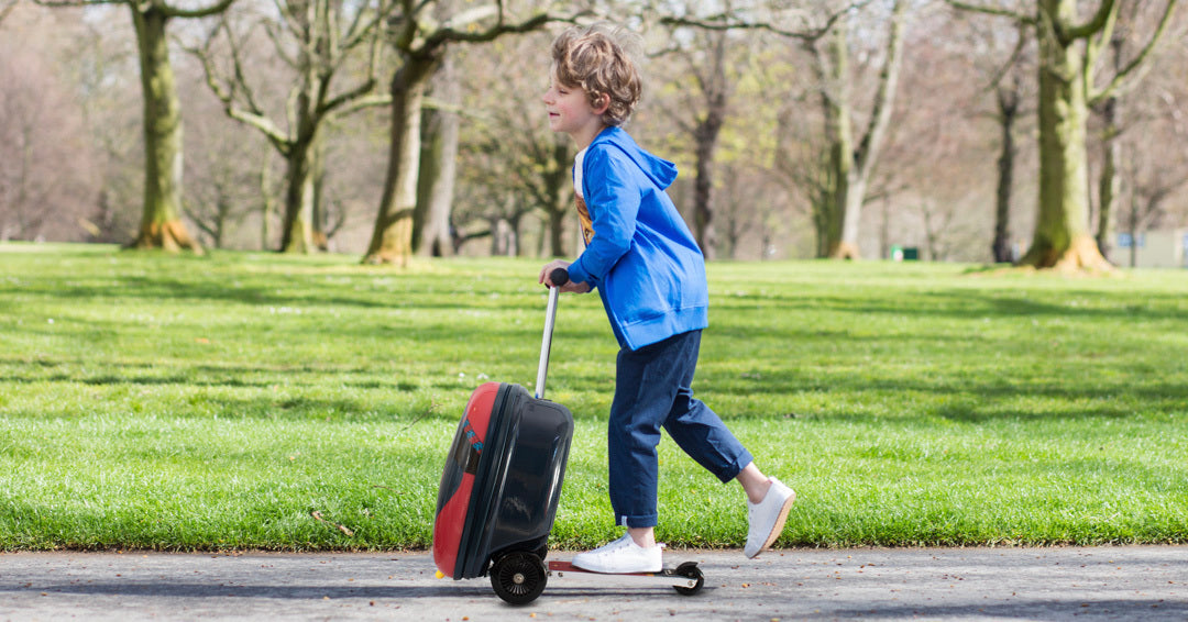 Boy riding scooter suitcase through park - fun travel luggage for kids