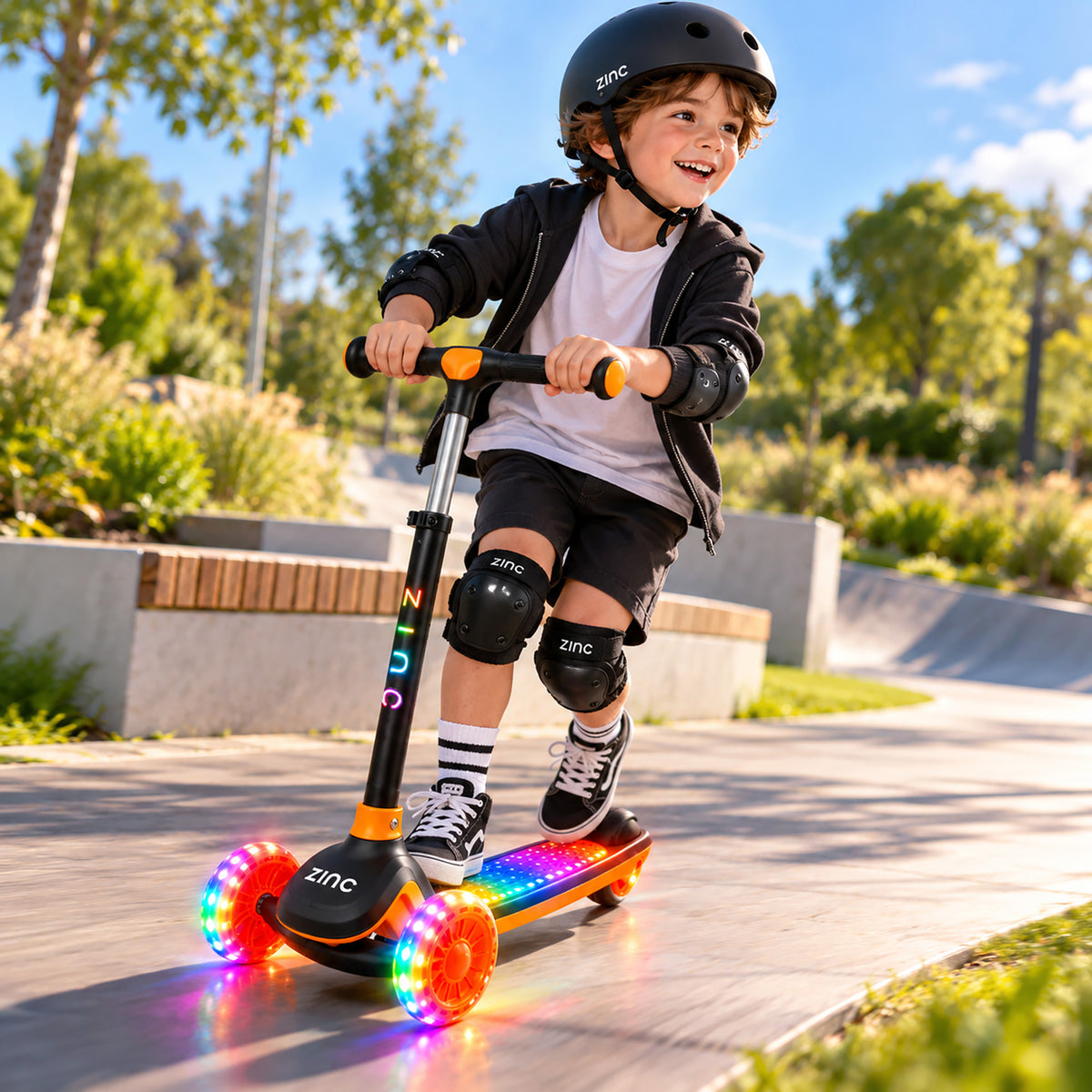 A child wearing a black helmet and protective gear rides a colourful LED light-up scooter on a sunny outdoor concrete path.
