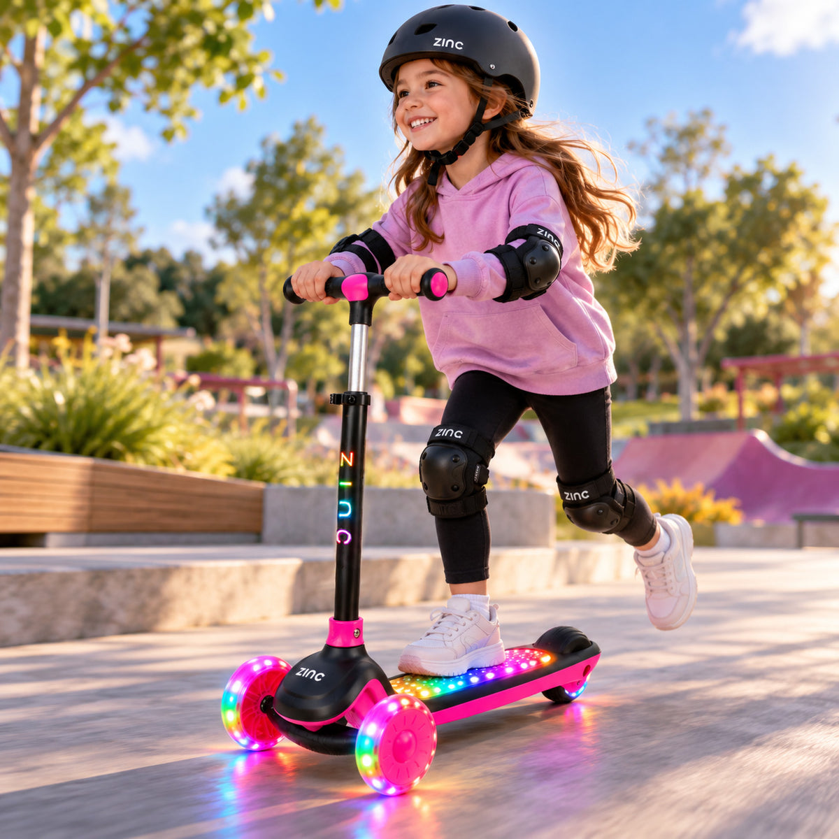 A young girl wearing a black helmet and protective gear rides a pink and black zinc scooter with glowing multicolor LED wheels in an outdoor park setting.