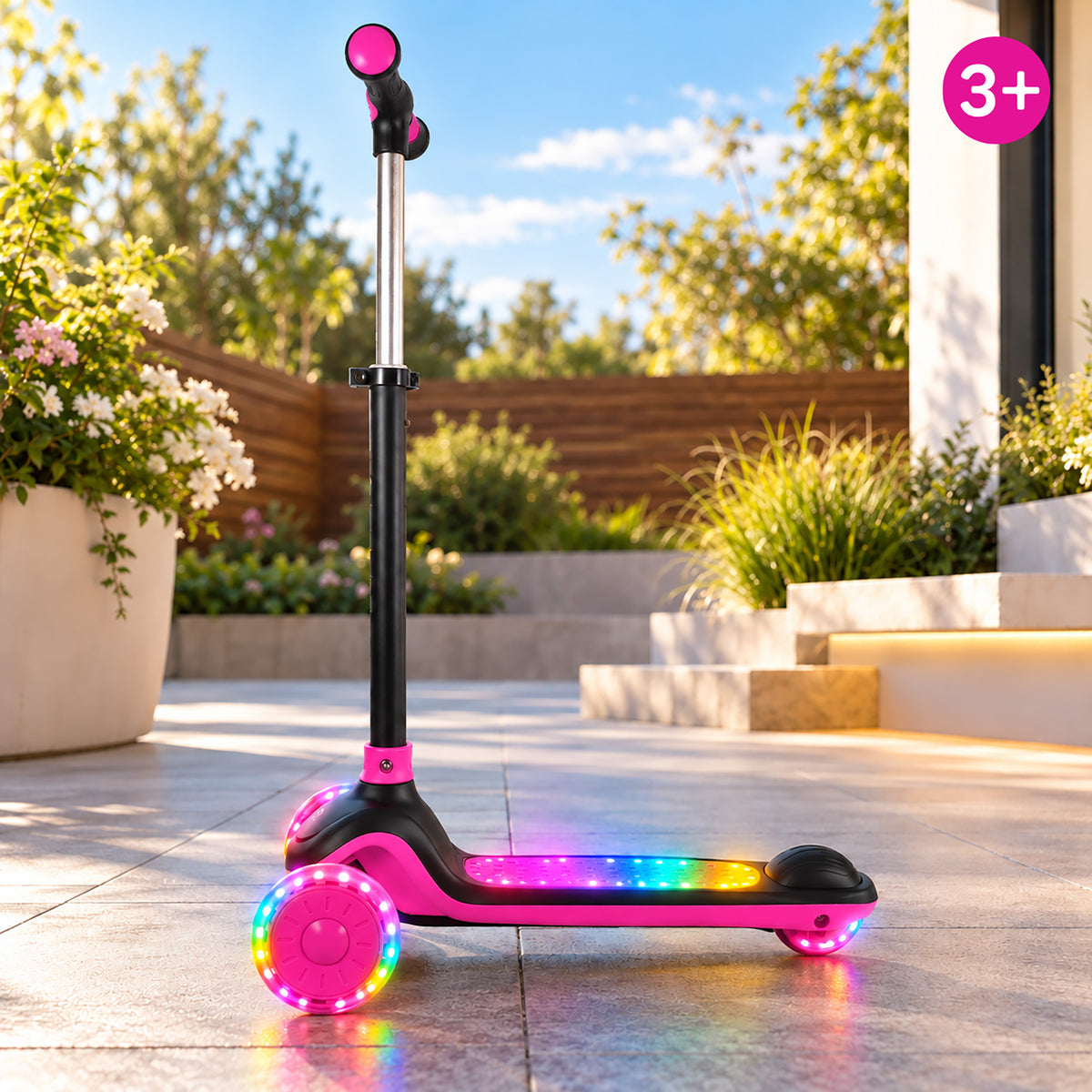 A pink and black three-wheel scooter with glowing multicolor LED lights on the deck and wheels, photographed outdoors on a paved patio with plants, steps, and a wooden fence in the background. A pink '3+' age badge appears in the top-right corner.