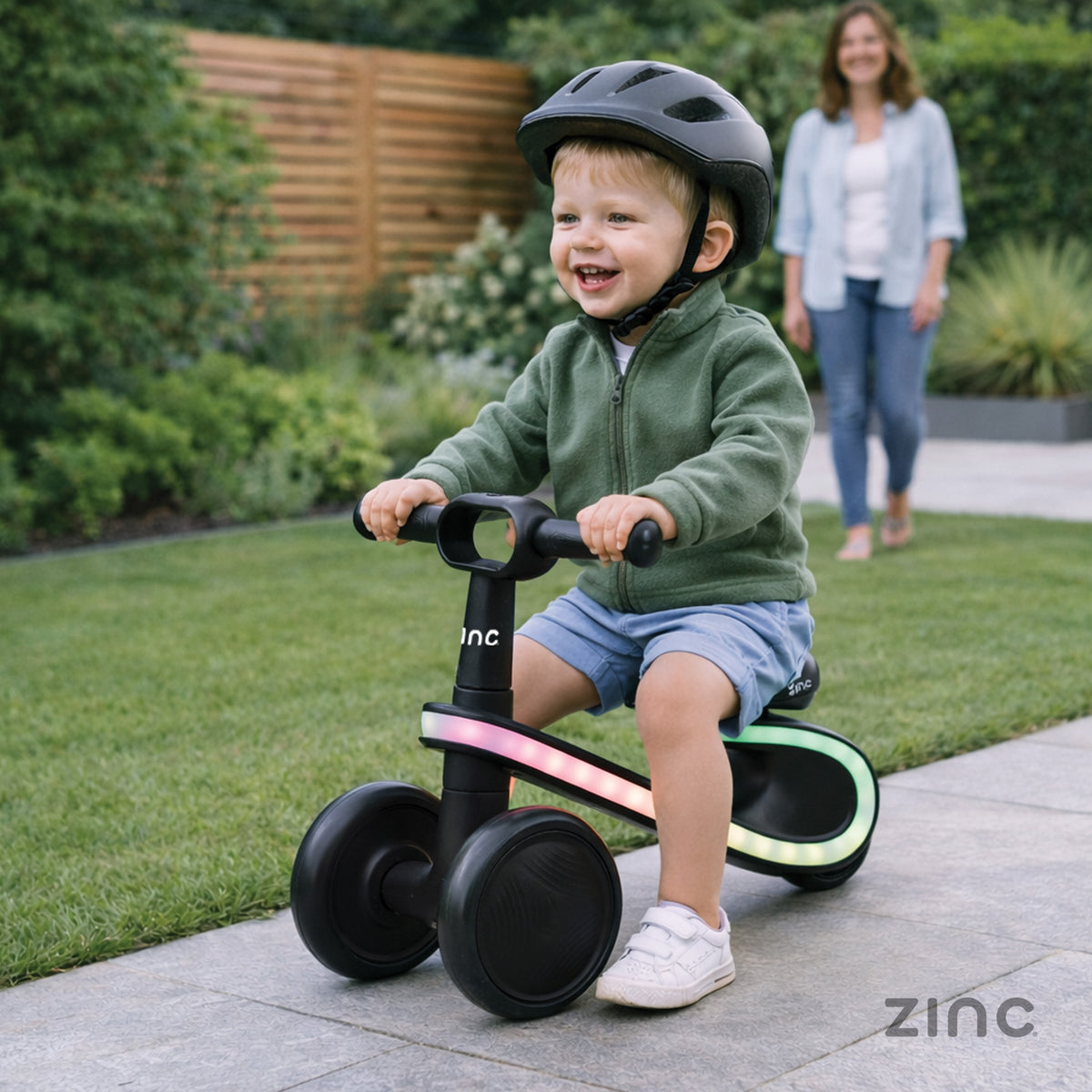 A toddler wearing a black helmet rides a black balance bike with colorful LED light accents on a paved path in a garden. A woman is visible in the background.