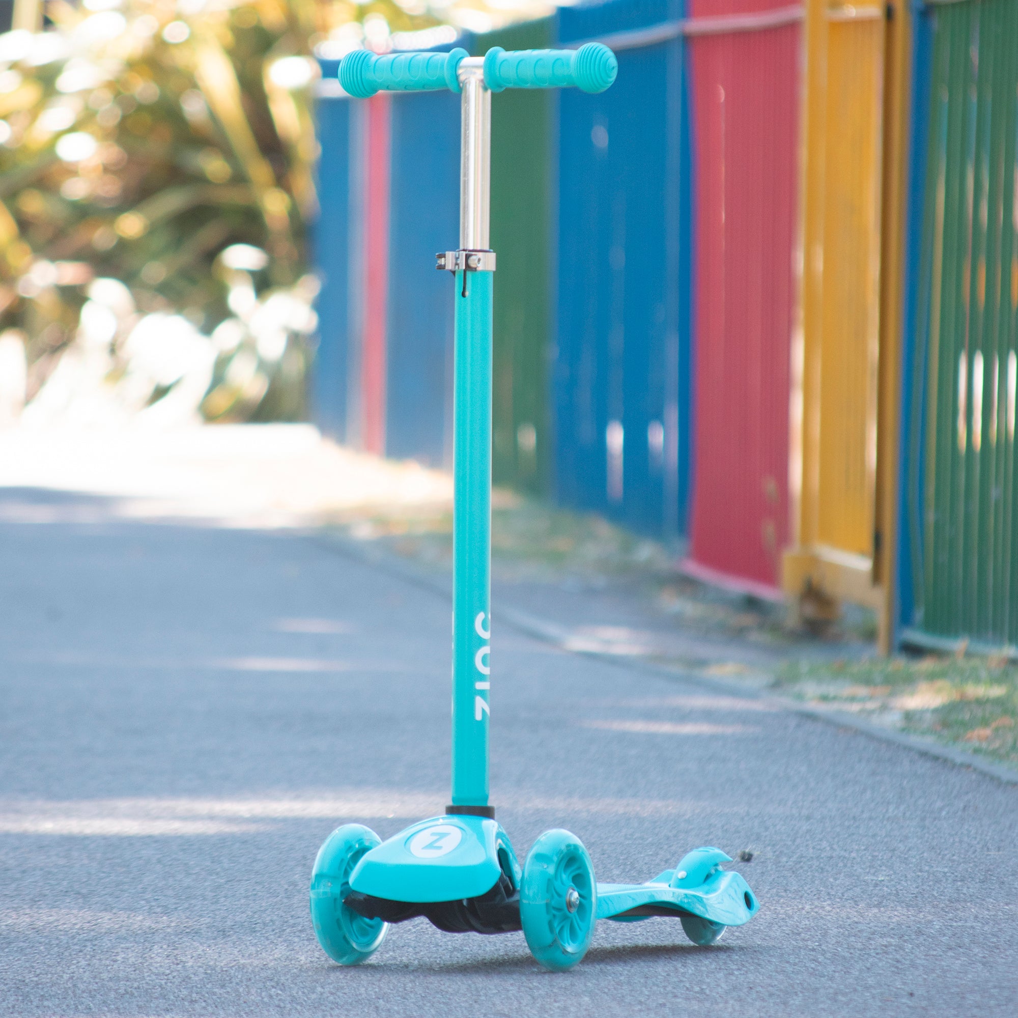 A pink Zinc three wheeled light up scooter with battery-free light up wheels, pictured on a pathway with colourful containers in the background.