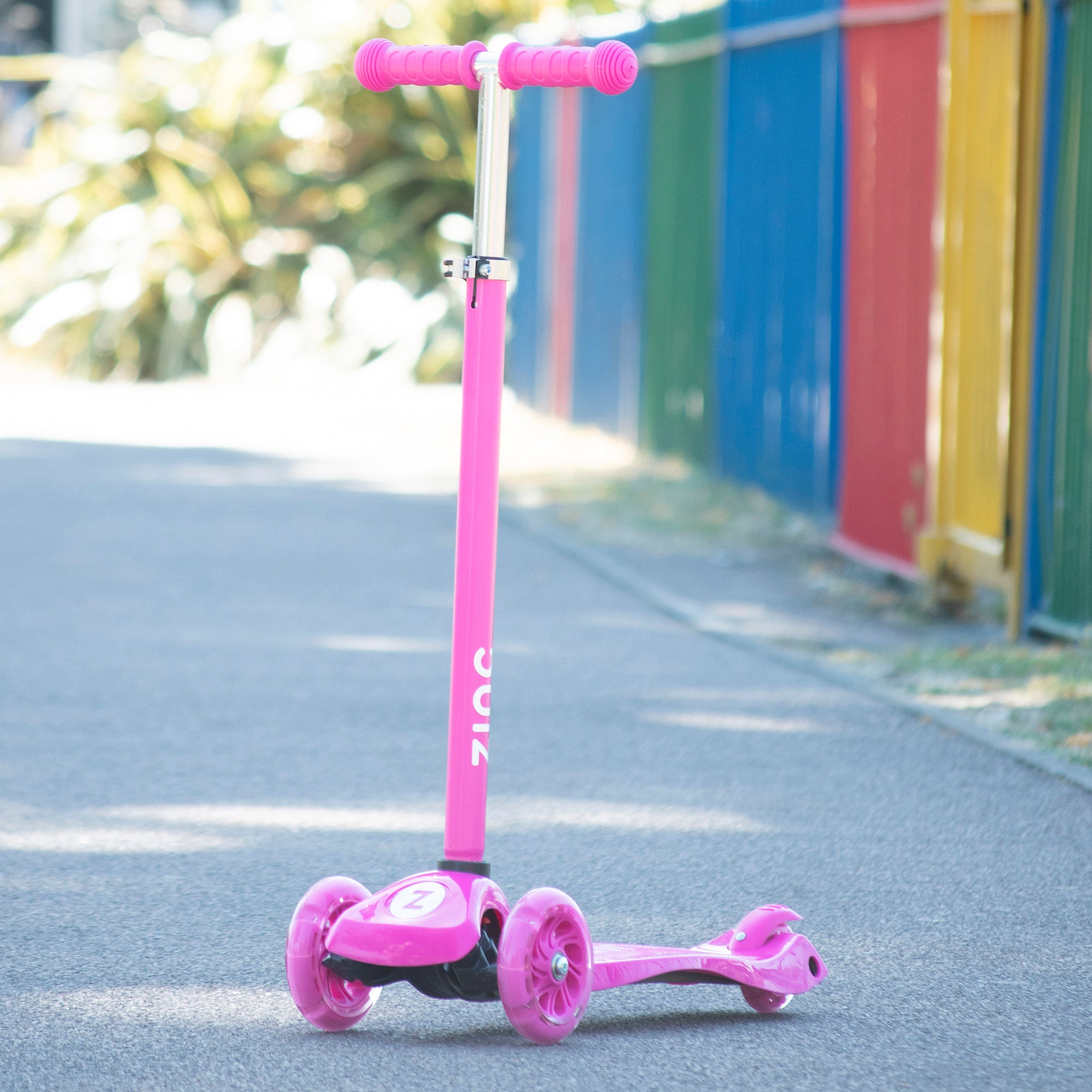 A pink Zinc three wheeled light up scooter with battery-free light up wheels, pictured on a pathway with colorful containers in the background.