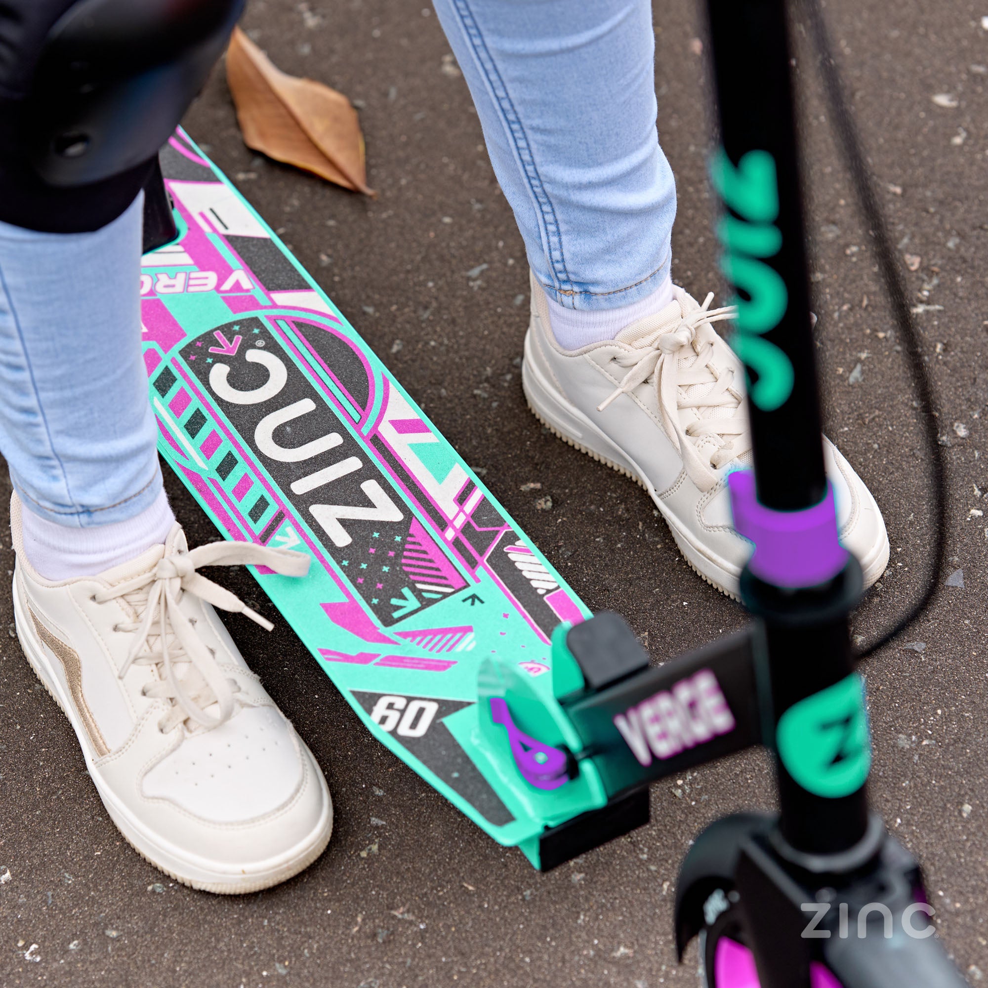 Person wearing white sneakers standing next to a green and pink scooter with 'Zinc' branding.