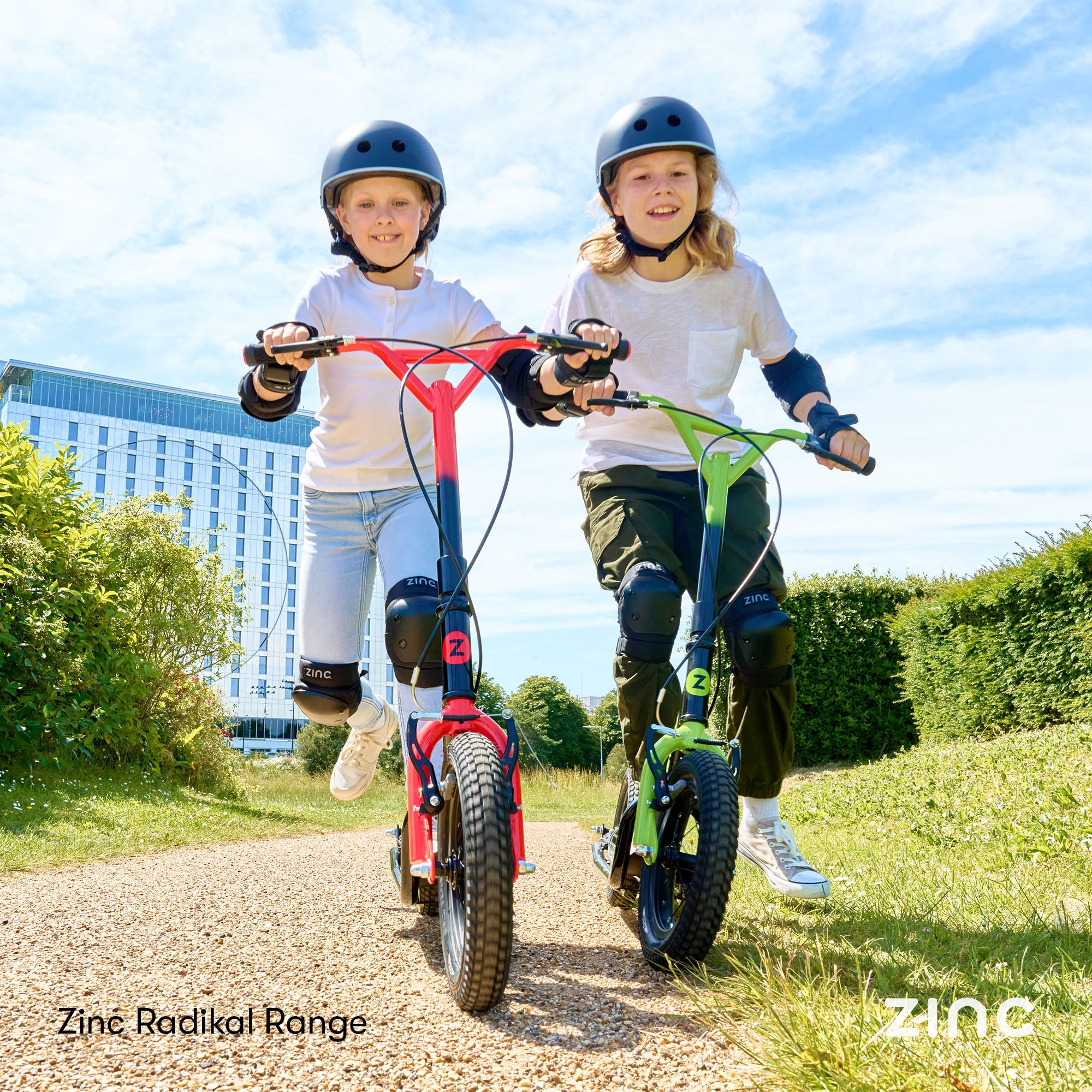 Boy and girl riding Zinc Radikal 12 inch BMX scooters outdoors in green and red colours