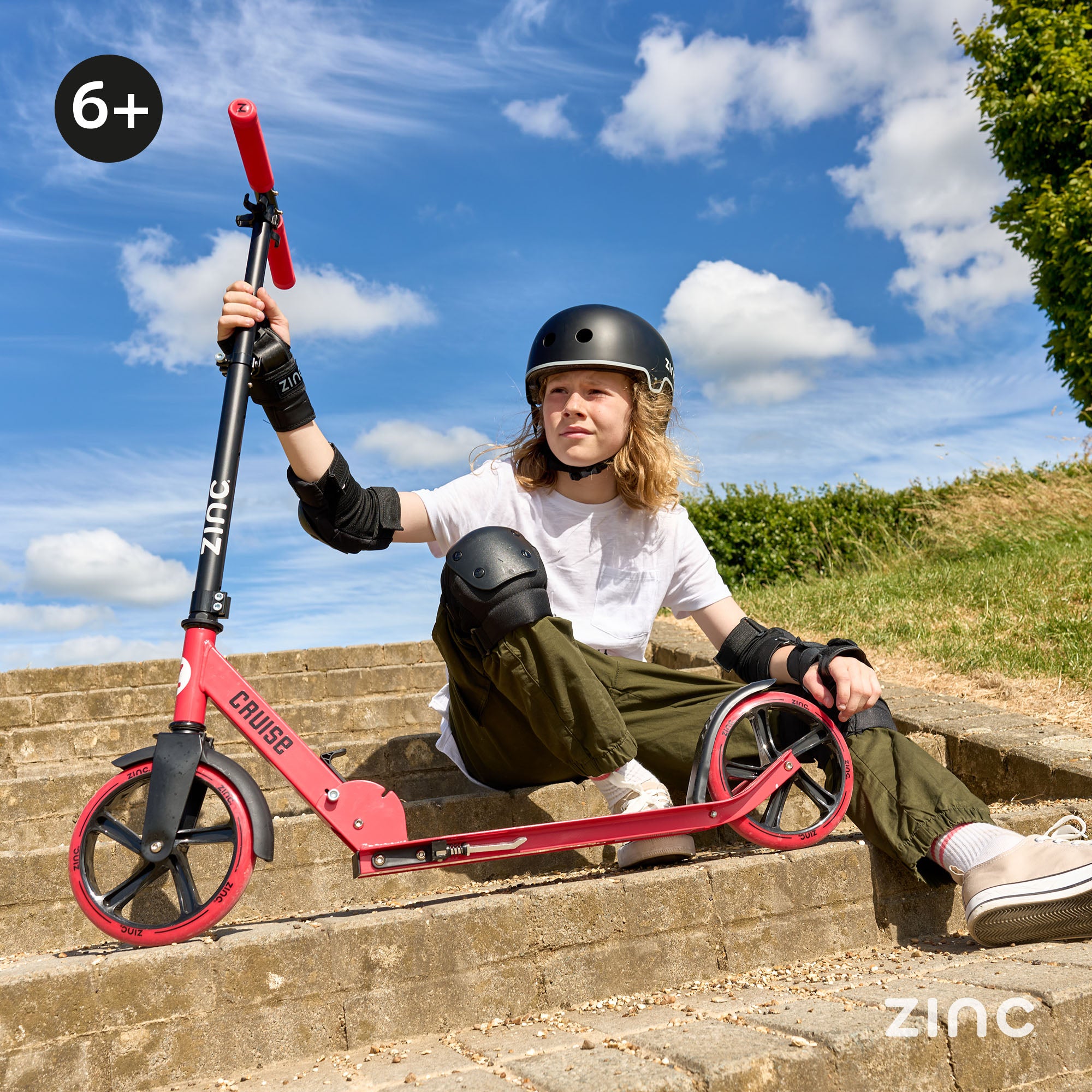 Boy sat with his Zinc Cruise big wheeled scooter in red with a blue sky background