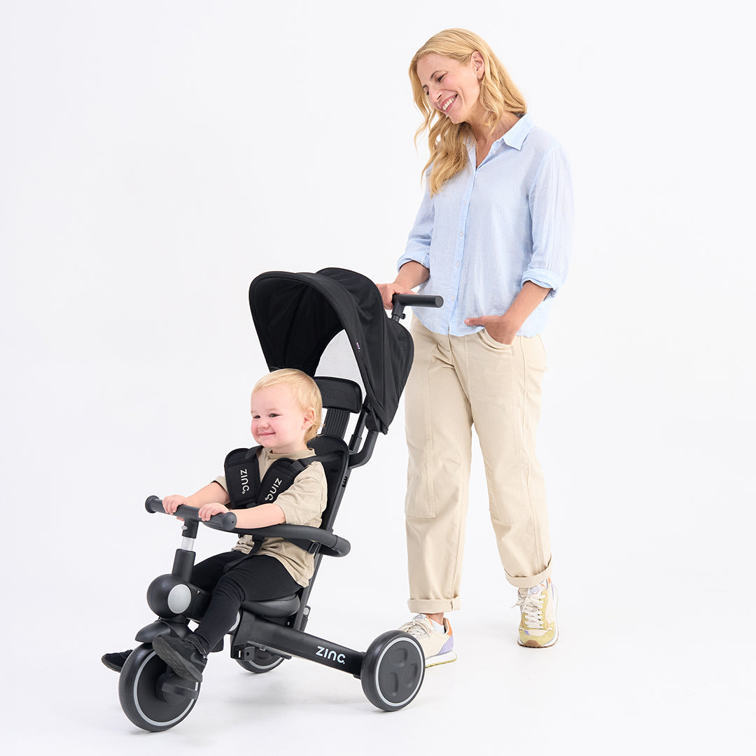 Child in a black multifunctional tricycle with a woman standing next to it on a white background.