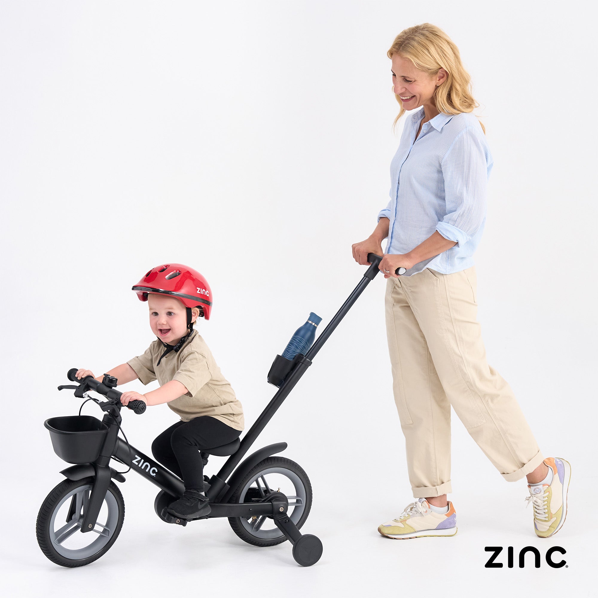 Woman pushing a child on a balance bike with a helmet, both smiling, on a white background with 'Zinc' branding.