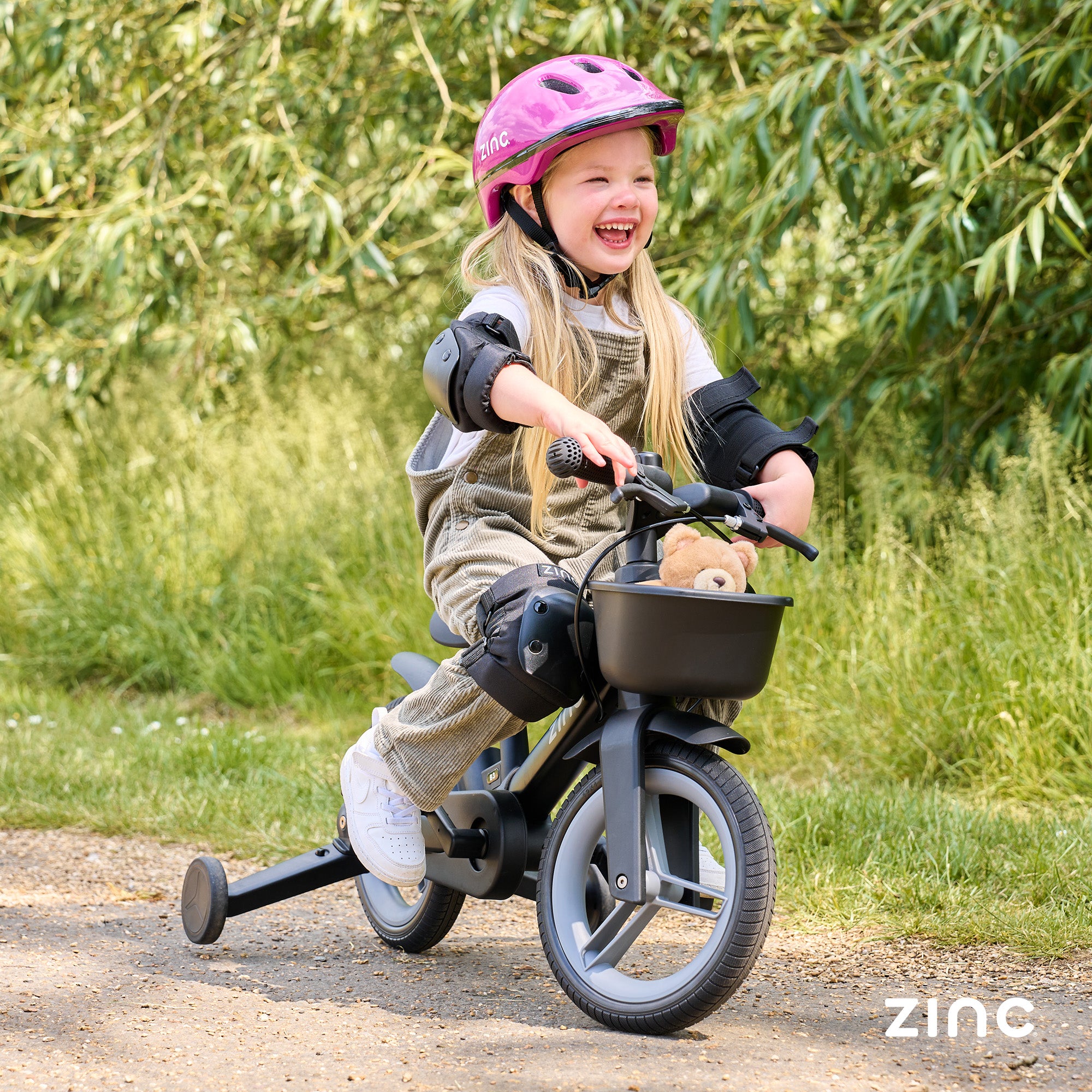 Child riding a tricycle with safety gear on a path surrounded by greenery, with 'zinc' branding.