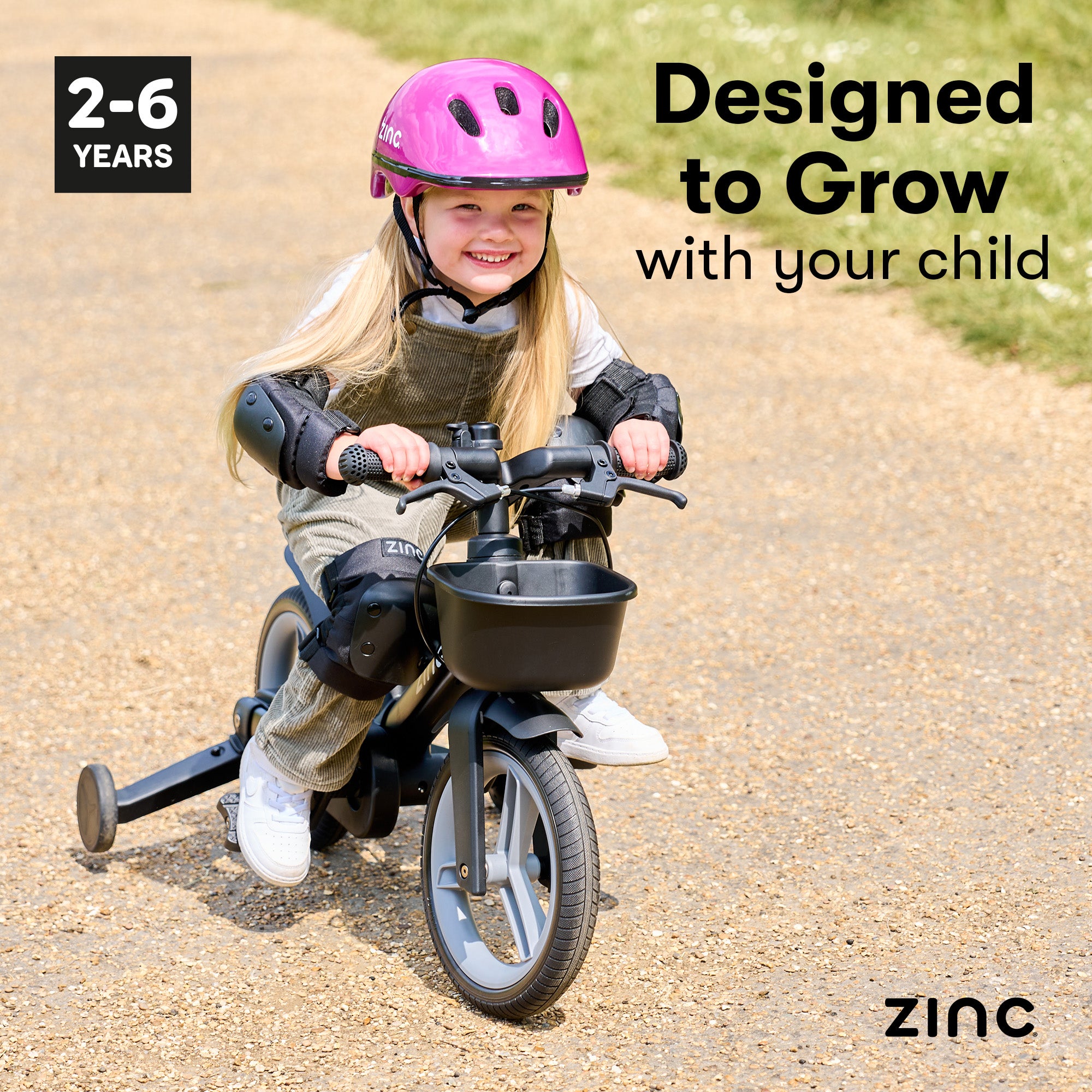 Child riding Zinc bike with stabilizers and safety gear, wearing a pink helmet, on a paved path with 'Zinc' branding.