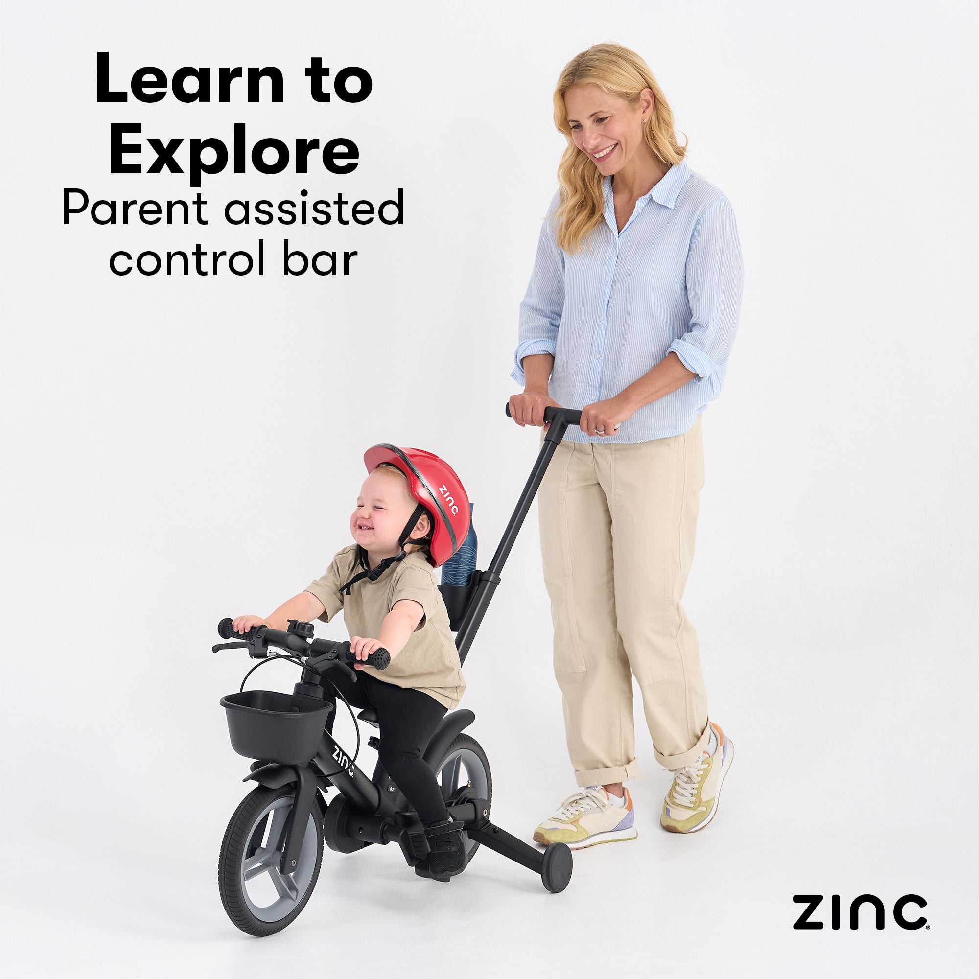 Woman pushing a child on a balance bike with a control bar, against a white background. Zinc brand visible.