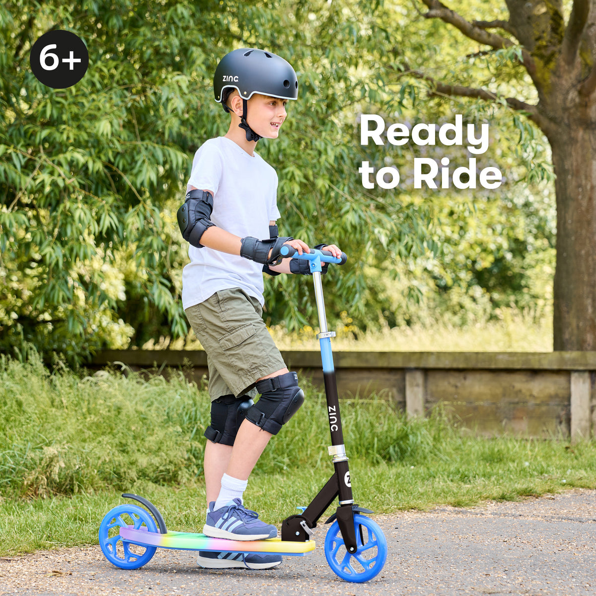 A child wearing a black helmet and protective pads rides a Zinc branded scooter outdoors on a paved path with green trees in the background. The scooter has a black frame, blue wheels and handlebar accents, and a rainbow-colored deck.