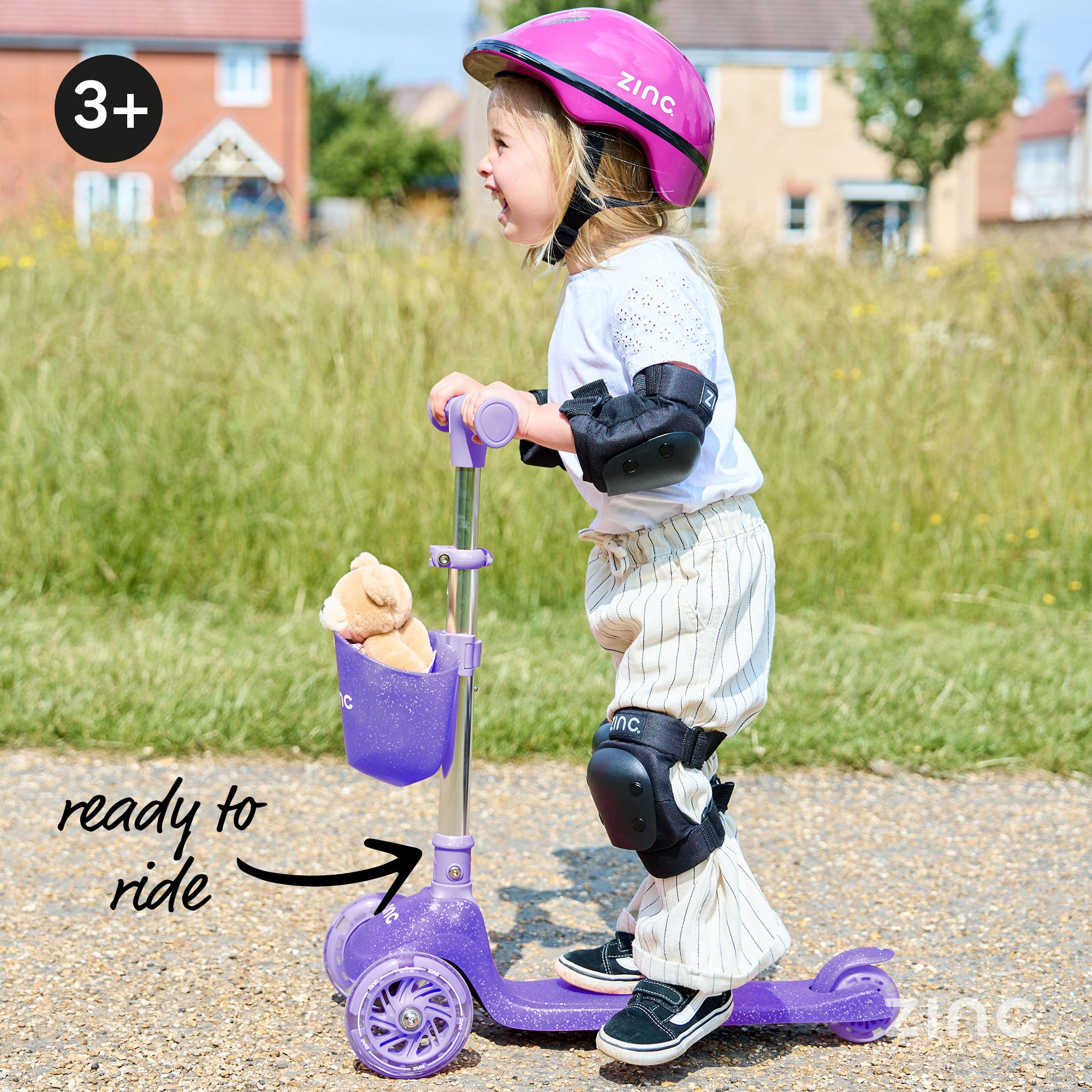 Child on a purple scooter with safety gear, standing on a path with grass and houses in the background.
