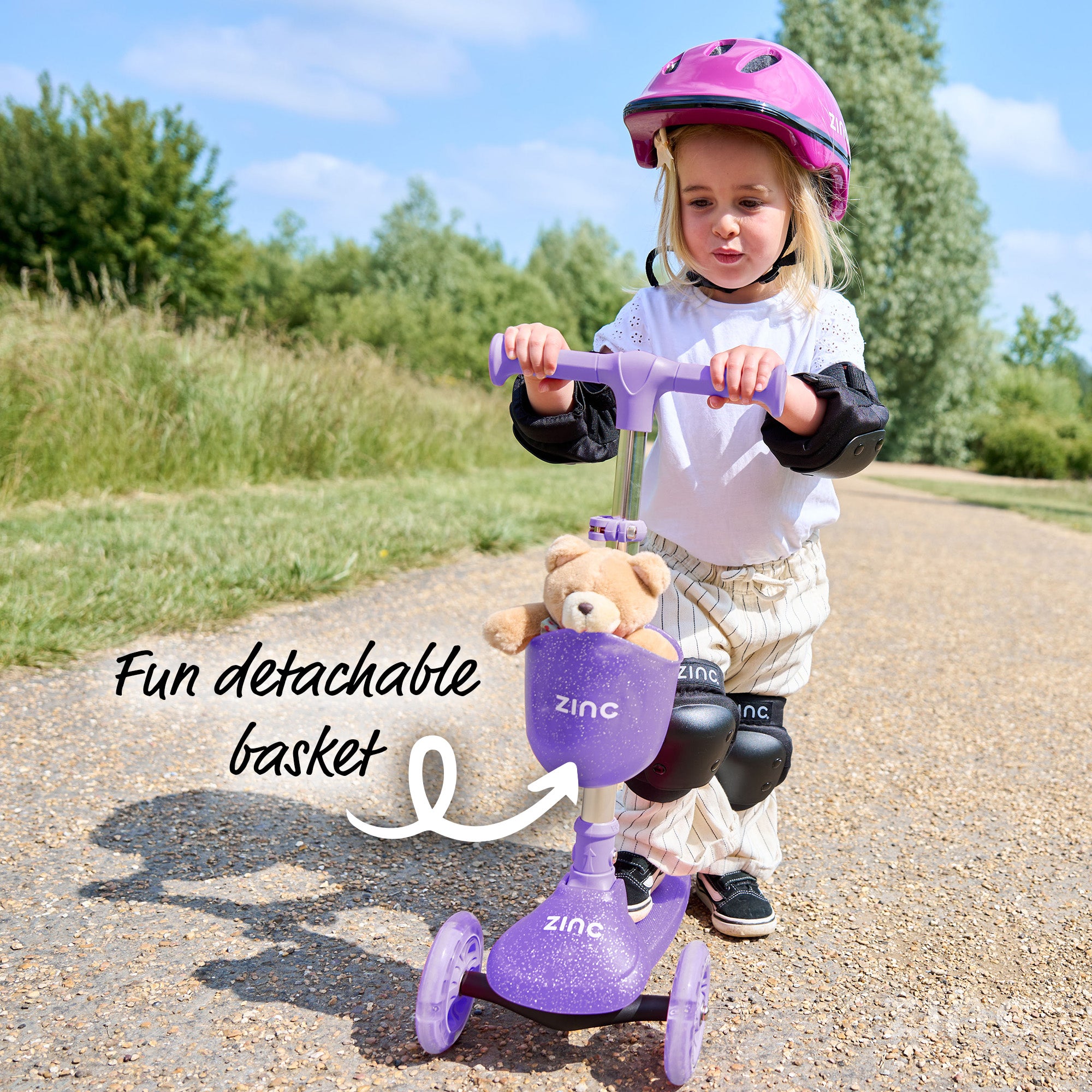Child riding a purple scooter with a detachable basket, wearing a helmet and knee pads, on a path with trees in the background.