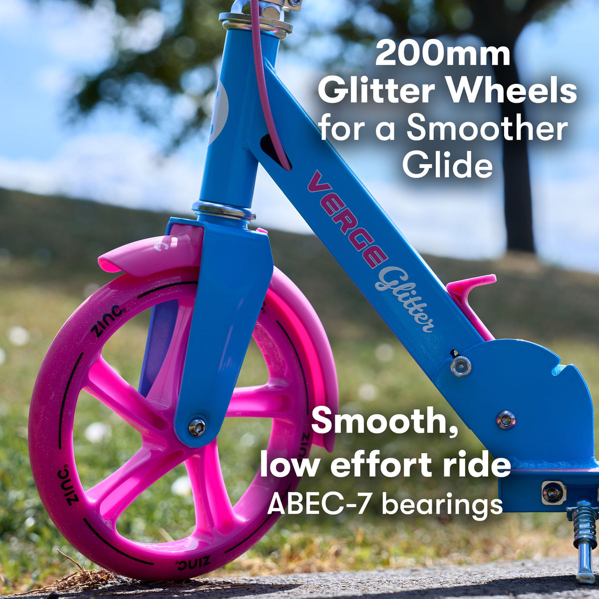 A close-up outdoor shot of a children's big wheel scooter wheel and frame. The frame is bright blue with pink wheel accents and visible text reading 'VERGE Glitter' and 'ZINC'. 