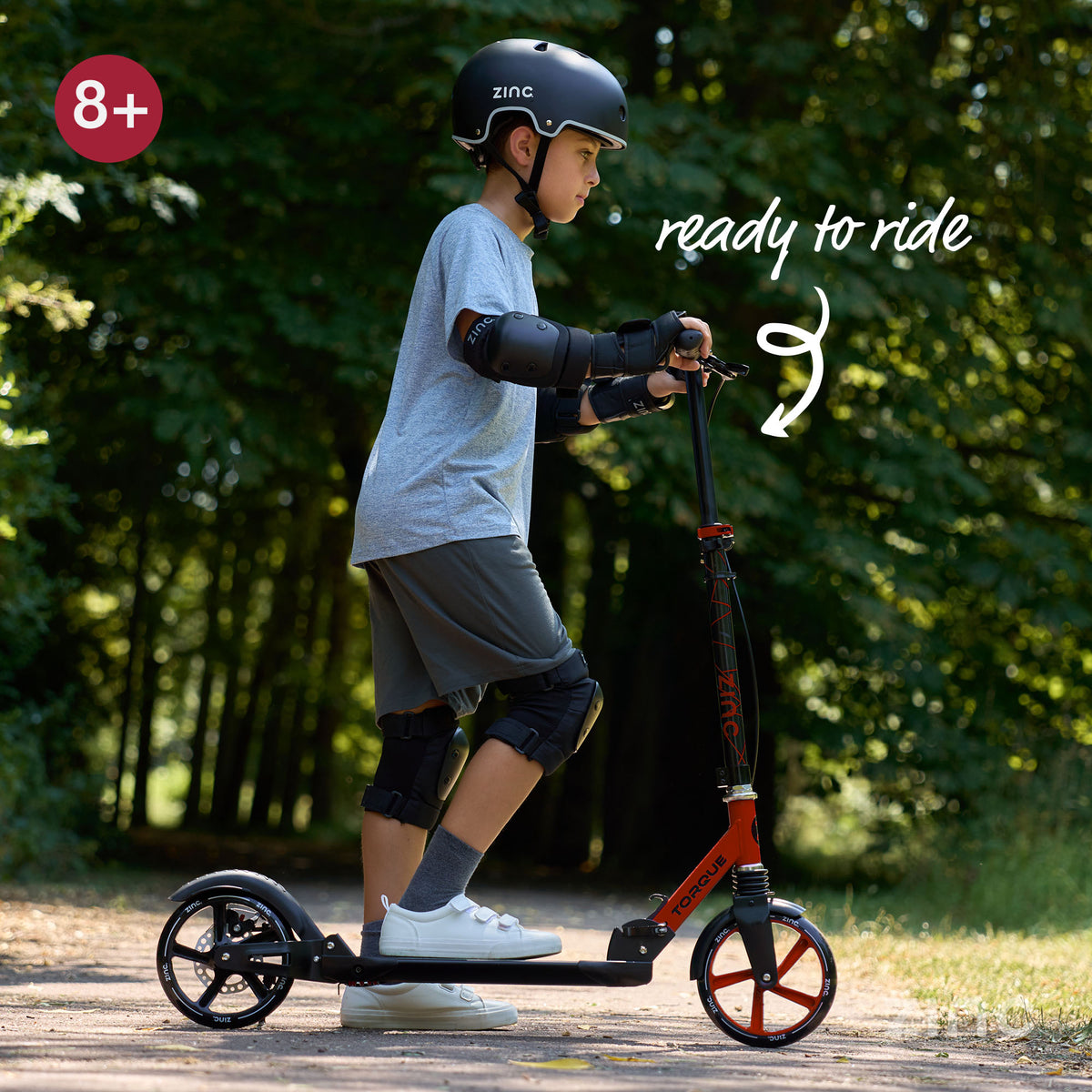 A child wearing a black helmet and protective pads rides a red and black big wheel kick scooter on a path in a wooded outdoor setting. White text reads 'ready to ride' with a curved arrow, and an '8+' age badge appears in the top left.