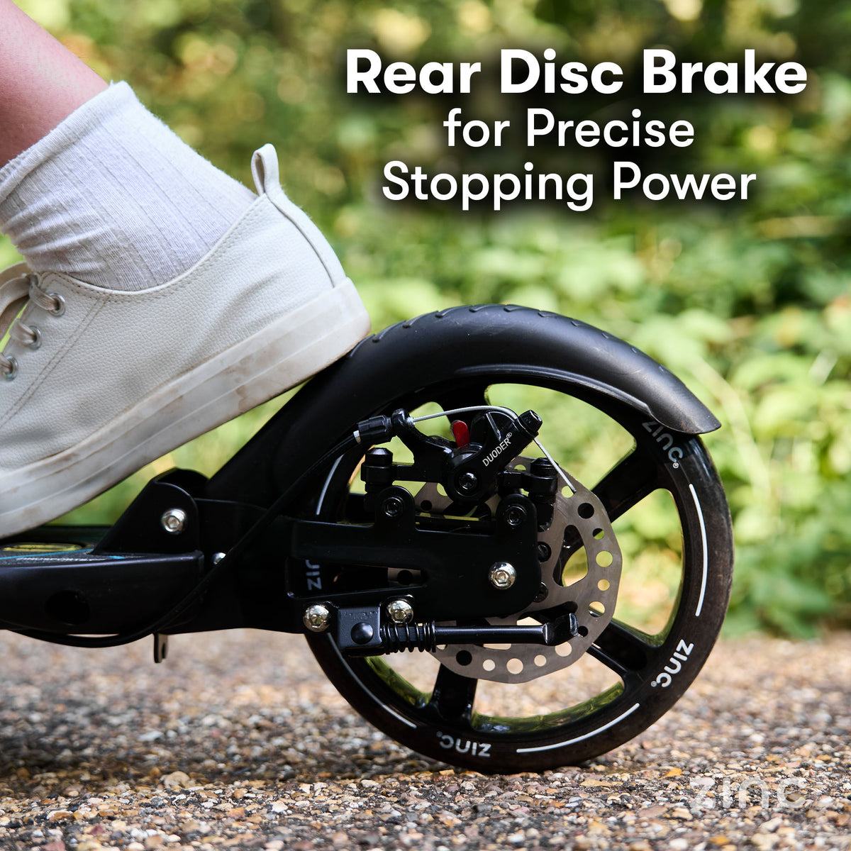 Close-up of a black scooter rear wheel with a disc brake, shown outdoors on gravel with a white shoe pressing against the brake. White text reads 'Rear Disc Brake for Precise Stopping Power.